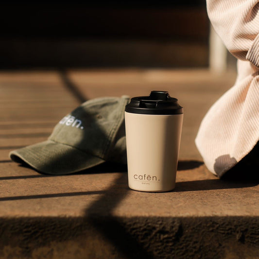 Disposable coffee cup with 'cafen' branding on a wooden surface, with a cap and green fabric in the background.